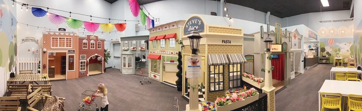 Inside an indoor playground built as a miniature town, with play storefronts and a child pushing a toy shopping cart